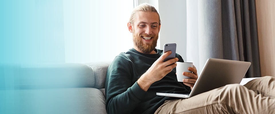 smiling man on couch with laptop looking at his mobile phone
